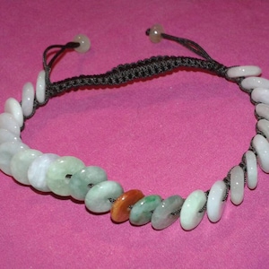 Op de afbeelding: Een jade armband met kleine, ronde, gepolijste jade stenen in tinten wit, groen en oranje. De stenen zijn geregen op een zwart geweven koord, met verstelbare uiteinden. De armband wordt getoond op een roze ondergrond.