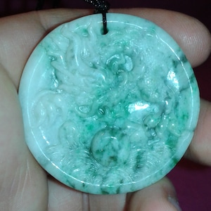 May include: Circular jade pendant featuring a carved dragon motif. The pendant's colours are a blend of white and green, with a black cord. The dragon is finely detailed, highlighting the craftsmanship of the piece.