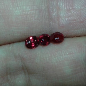 May include: Three small, round, faceted gemstones in a vibrant red hue. The gems are displayed on a light-toned background, showcasing their polished surfaces and internal brilliance. The gems are likely rubies or garnets.
