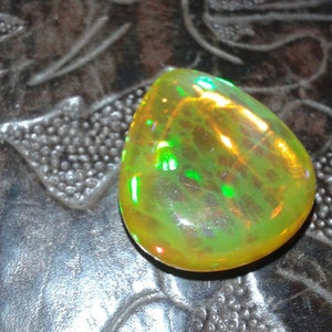 May include: A pear-shaped, polished opal gemstone. The opal displays a vibrant play-of-color, with flashes of green and yellow hues. The stone is set against a dark, textured surface, highlighting its iridescent qualities.