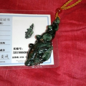 May include: A dark green jade pendant with a carved design, attached to a gold-colored cord. A smaller jade piece and a certificate are also visible. The pendant is on a red background.