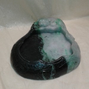 May include: A carved jade bowl with a dark green exterior and a lighter green and white interior. The bowl has a rounded shape with carved details and a smooth, polished finish. The bowl is likely used for decorative purposes.