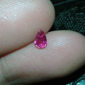 May include: A small, pear-shaped, faceted gemstone in a vibrant pink hue. The gem is held between two fingers, showcasing its size and brilliance. The background is a dark, textured surface.