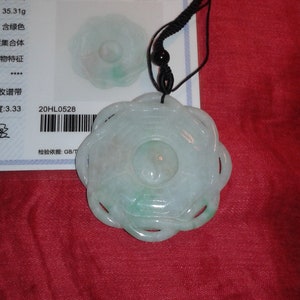 May include: A carved jade pendant with a floral design, featuring a central circular element and openwork petals. The pendant is suspended from a black cord. The jade has a pale green hue. A certificate is visible in the background.