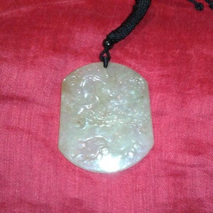 Puede incluir: Colgante de jade verde pálido con cordón negro. El colgante tiene forma hexagonal irregular con detalles tallados. El fondo es de tela roja.