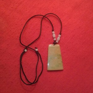Puede incluir: Un collar con colgante de jade con un colgante rectangular verde claro y amarillo. El colgante está suspendido de un collar de cordón negro con cuentas blancas. El fondo es de color rojo liso.