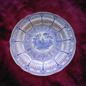 May include: A blue and white decorative plate with a calendar for 2001. The plate has a detailed border with months and dates, and a central illustration of a pastoral scene. The plate is round with a scalloped edge, typical of fine china.