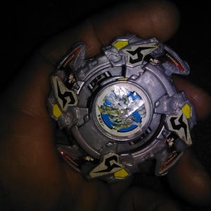 May include: A silver and grey spinning top toy with yellow and red details. The centre has a circular design with a blue and green landscape. The toy is held in a hand against a dark background.