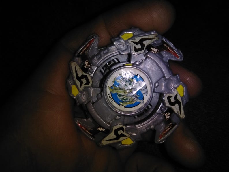 May include: A silver and grey spinning top toy with yellow and red details. The centre has a circular design with a blue and green landscape. The toy is held in a hand against a dark background.
