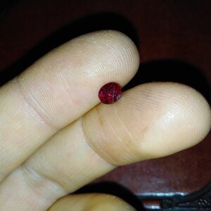 Op de afbeelding: Een kleine, ovaalvormige, dieprode edelsteen wordt tussen twee vingers gehouden. De edelsteen heeft een enigszins ruw oppervlak en lijkt gefacetteerd te zijn. De achtergrond is een donker, wazig oppervlak.