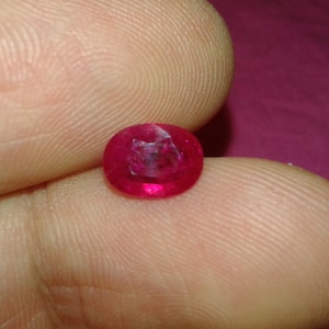 May include: A round, faceted ruby gemstone, displaying a vibrant red hue. The gemstone is held between two fingers, showcasing its polished surface and internal clarity. The ruby's color is a deep, saturated red, typical of high-quality gemstones.