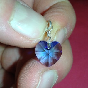 May include: A faceted, heart-shaped pendant with a silver-colored wire loop. The pendant is a deep blue color with iridescent highlights. The heart is held between fingers against a pink background. Jewelry.