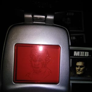 以下が含まれることがあります： 赤いスクリーンにキャラクターのスケッチが表示された、銀色の小道具デバイス。 デバイスは長方形で、ヒンジ付きのトップが付いています。 画像には「MIB」というテキストのカードも表示されています。