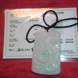 Puede incluir: Un colgante rectangular de jade con un diseño tallado, suspendido de un cordón negro. El colgante es principalmente blanco con acentos verdes. Un certificado con texto chino es visible en el fondo.