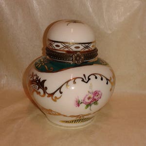 May include: A white and green porcelain jar with a lid, featuring floral and gold accents. The jar has a decorative metal band around the middle and a floral design on the front. The lid is white with a small gold design.