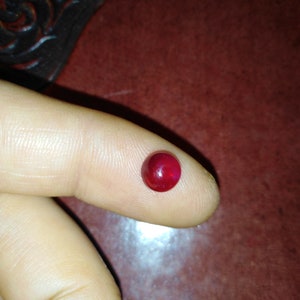 Pode incluir: Uma pequena gema redonda, polida, vermelho escuro, possivelmente um rubi, repousa sobre um dedo. A gema é lisa e reflete a luz, mostrando sua cor rica. O fundo é uma superfície marrom avermelhada escura.