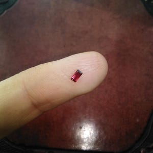 May include: A small, rectangular, deep red gemstone, possibly a ruby or garnet, is displayed on a fingertip. The gem has a polished, faceted appearance, catching the light. The background is a dark, textured surface.