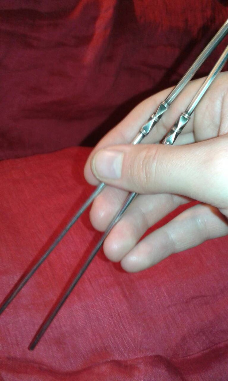 May include: A pair of silver chopsticks with decorative diamond-shaped accents. The chopsticks are held against a red fabric background, showcasing their sleek design and potential for Asian-inspired dining.