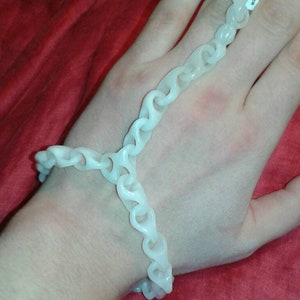 May include: A white, chain-link bracelet and ring set. The bracelet wraps around the wrist, and a chain extends to the ring on the adjacent finger. The jewelry is made of a light-colored, possibly plastic material.