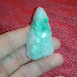 Pode incluir: Um pingente de jade esculpido em forma de Buda. O pingente é de cor verde claro com detalhes em verde escuro. A escultura tem características suaves e arredondadas e é segurada em uma mão.