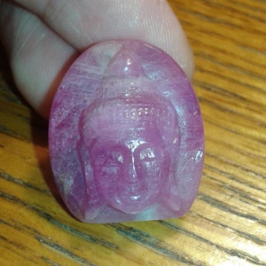 May include: A carved, oval-shaped, pink gemstone featuring a detailed depiction of a Buddha's face. The carving showcases intricate details of the Buddha's serene expression and headdress. The gemstone is held against a wooden background.