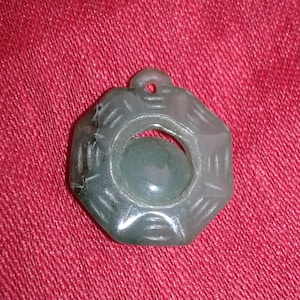 May include: A jade pendant with an unusual octagonal shape and a central circular design. The pendant is a muted green colour and has a small hole at the top for a chain. The background is a textured red fabric.