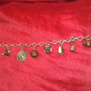 Puede incluir: Una pulsera de dijes plateada con varios dijes pequeños y detallados. Los dijes incluyen un estribo, un pájaro, un plato decorativo, una tetera, una caja pequeña, una campana y dos dijes rectangulares. La pulsera se muestra sobre una tela roja.