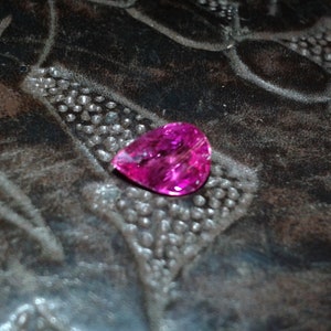May include: A vibrant, heart-shaped, fuchsia gemstone rests on a textured, dark brown surface. The gemstone's facets catch the light, displaying its brilliance. The background has a pattern of small, raised dots and curved lines.