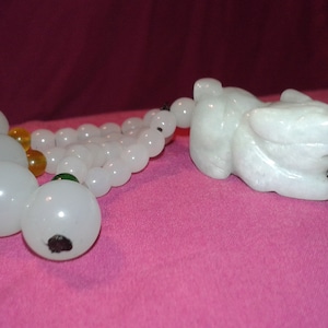 Puede incluir: Un colgante de jade blanco con una figura de animal tallada y una cadena de cuentas blancas y ámbar. El colgante se muestra sobre una superficie rosa.