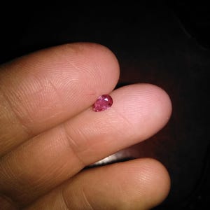 Può includere: Una piccola gemma rosa, sfaccettata, a forma di goccia. La gemma è tenuta tra due dita su uno sfondo scuro. Le sfaccettature della gemma catturano la luce, creando un effetto scintillante. La gemma è probabilmente un rubino o uno zaffiro rosa.