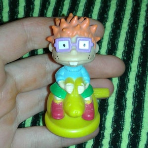 Puede incluir: Una pequeña figura de juguete de plástico de Chuckie Finster de la serie animada Rugrats. La figura tiene pelo naranja, gafas moradas y lleva una camisa azul, pantalones verdes y zapatos rojos. El juguete está montado en un caballito balancín amarillo.