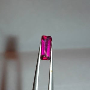 May include: A rectangular, vibrant pink gemstone is held by silver tweezers. The gem has a polished surface and a clear, transparent appearance. The gemstone's color is a deep, saturated pink, with a slight variation in tone.