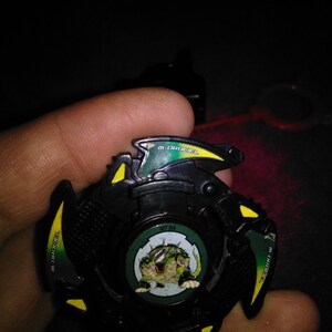 May include: A black and green toy with yellow accents, featuring a circular design with a cartoon creature. The toy has the text "M-Drager" and "MT-03" on it. A red pull string is visible.