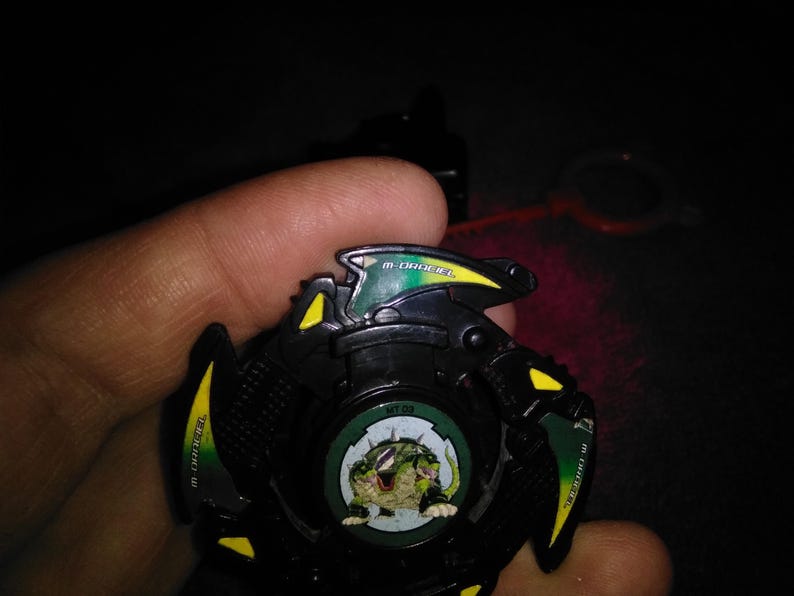 May include: A black and green toy with yellow accents, featuring a circular design with a cartoon creature. The toy has the text "M-Drager" and "MT-03" on it. A red pull string is visible.
