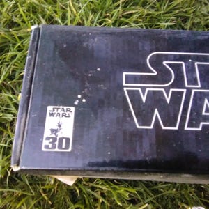 Peut inclure: Une boîte noire avec le logo Star Wars en blanc. La boîte présente également le texte "STAR WARS" en grandes lettres blanches et un logo plus petit avec le texte "STAR WARS" et le chiffre "30".