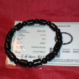 Puede incluir: Una pulsera de cuentas negras con cuentas cilíndricas y esféricas. La pulsera se muestra sobre una superficie roja junto a una tarjeta blanca con texto y un diagrama de la pulsera. El peso total es de 22,5 g.