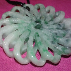 May include: A jade pendant with a floral design. The pendant is carved with a series of curved, petal-like shapes, creating a circular pattern. The jade is a mix of white and green hues, with a black cord for wearing.