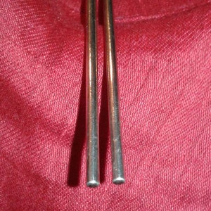 May include: Two slender, metallic chopsticks with rounded tips are positioned vertically against a textured, deep red background. The chopsticks have a polished, reflective surface, suggesting a dining or culinary theme.