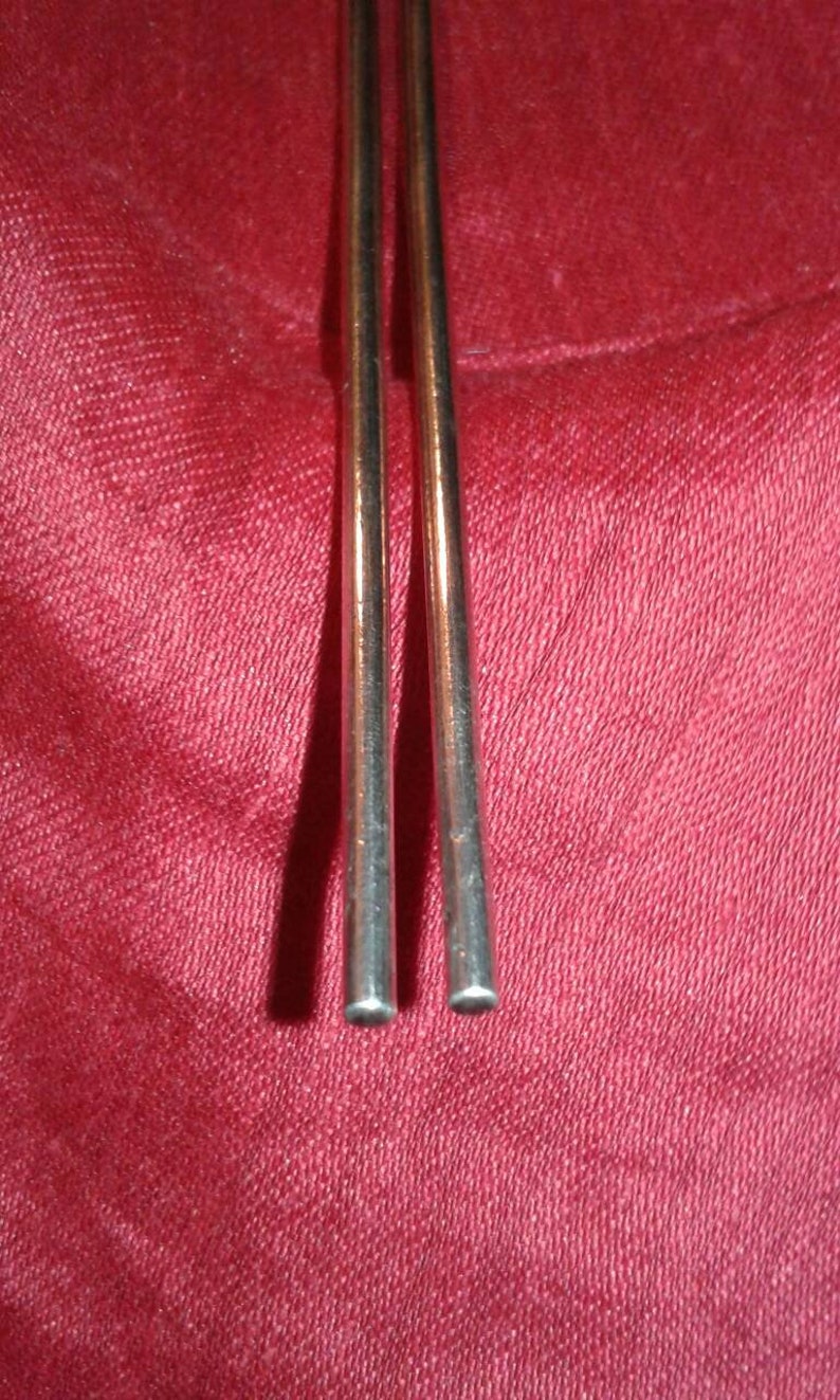 May include: Two slender, metallic chopsticks with rounded tips are positioned vertically against a textured, deep red background. The chopsticks have a polished, reflective surface, suggesting a dining or culinary theme.