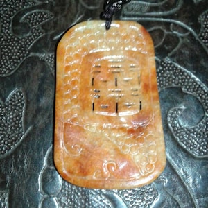 Könnte beinhalten: Eine rechteckige Anhängerhalskette mit einem geschnitzten Design. Der Anhänger hat eine warme orange-braune Farbe mit einem geschnitzten Muster und einem zentralen Design. Die Halskette hat eine schwarze Kordel und weiße Perlen.