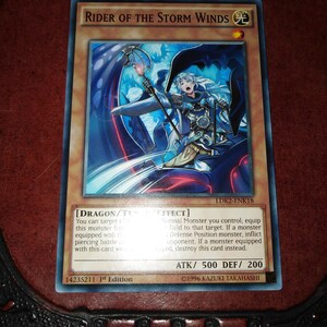以下が含まれることがあります： 「Rider of the Storm Winds」と書かれたトレーディングカード。青と白のイラストで、ドラゴンに似た生き物にまたがるキャラクターが描かれています。カードには属性と効果を説明するテキストがあります。ATKは500、DEFは200です。