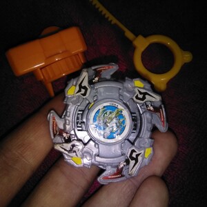 May include: A silver and grey toy with yellow and red details, featuring a central graphic and the text "GER4". Includes an orange launcher and a yellow ripcord. A children's toy.