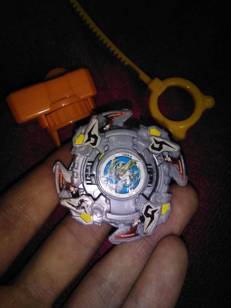 May include: A silver and grey toy with yellow and red details, featuring a central graphic and the text "GER4". Includes an orange launcher and a yellow ripcord. A children's toy.