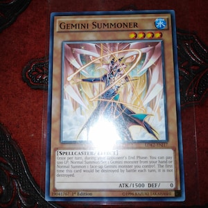 以下が含まれることがあります： 「GEMINI SUMMONER」と題された遊戯王トレーディングカード。青と黒のローブを着たキャラクターが杖を持ち、幾何学模様の背景に配置されています。カードには水の属性と初版のマークがあります。