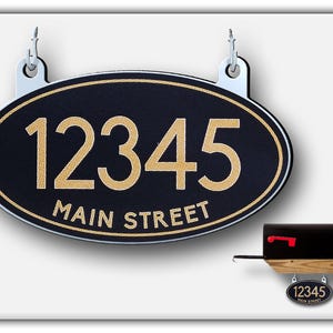 May include: Black oval-shaped house number sign with gold lettering. The sign reads "12345 MAIN STREET" and is hanging from two silver hooks. A smaller version of the sign is attached to a wooden mailbox.