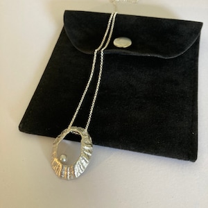 Puede incluir: Un collar plateado con un colgante ovalado y una pequeña perla. El colgante tiene un diseño texturizado y orgánico. El collar se muestra en una bolsa de terciopelo negro con cierre de botón a presión.