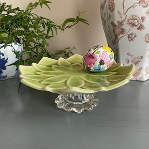 May include: A light green ceramic serving dish with a flower petal design, resting on a clear, ornate glass base. An egg decorated with colorful flowers sits on the dish. A potted plant and a decorative vase are in the background.