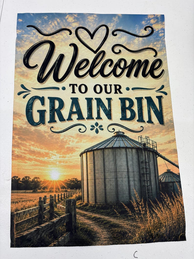 May include: A rectangular garden flag featuring a sunset landscape with a metal grain bin, a wooden fence, and a dirt path. The text "Welcome TO OUR GRAIN BIN" is printed in black and blue lettering at the top, accompanied by a heart graphic.