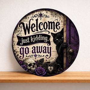 Può includere: Un cartello circolare in legno con un gatto nero che sbuca da una porta viola, un teschio e rose viola. Il testo Welcome just kidding, go away è scritto in nero. Lo sfondo include ragnatele e una tazza fumante con un cuore disegnato.