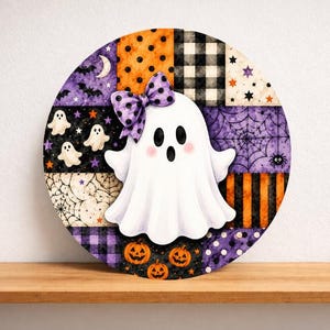 May include: A circular wall sign featuring a patchwork design of Halloween patterns including orange pumpkins, black and white buffalo plaid, purple spiderwebs, and bats. A central white ghost with a purple polka dot bow sits in the middle.